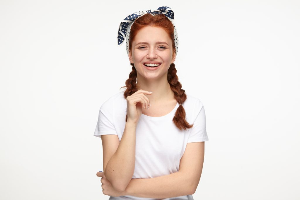 portrait of young ginger female standing over white studio background keeps hands folded and smiles broadly