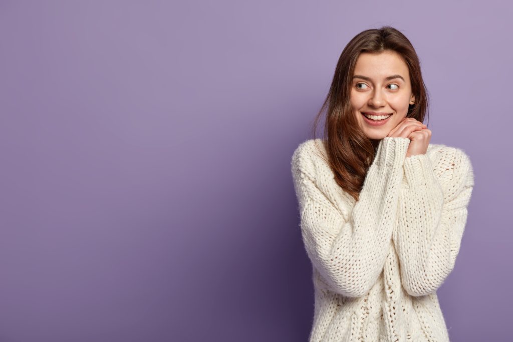 cheerful caucasian girl keeps hands together near face, looks positively aside, has no make up, healthy skin, wears white sweater, stands over purple background with blank space for your promotion